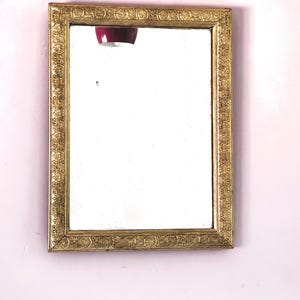 May include: A rectangular mirror with a gold-colored ornate frame. The frame features detailed floral and scrollwork patterns. The mirror is hanging on a light pink wall.