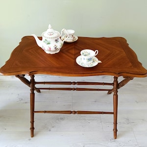 Peut inclure: Une table à thé en bois brun foncé avec un bord décoratif. La table est ornée d'une théière à fleurs, de deux tasses et de soucoupes. Les pieds de la table sont fins et comportent des supports horizontaux. La table mesure environ 75 cm de long.