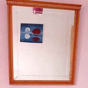Pode incluir: Um espelho retangular com moldura de madeira. A moldura é de cor castanho quente com uma borda branca decorativa. O espelho reflete uma pequena pintura de uma árvore vermelha e duas luas. Uma abajur rosa é visível acima do espelho.