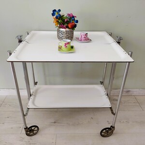 May include: A vintage white serving cart with a lower shelf, supported by silver-colored metal legs and small wheels. The cart has a white tray on top with a floral teacup and saucer, and a vase of flowers.