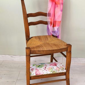 Peut inclure: Chaise en bois avec assise en paille tressée et coussin fleuri. Une écharpe transparente aux motifs géométriques roses, rouges et violets est suspendue au dossier. Design classique et vintage.