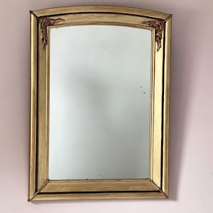 Può includere: Uno specchio da parete rettangolare con una cornice color oro e un bordo superiore arcuato. La cornice presenta decorazioni intagliate a forma di foglia negli angoli superiori. La superficie in vetro è chiara e riflette la luce.