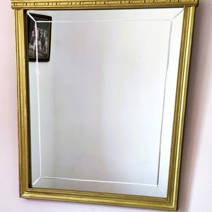 May include: A rectangular gold-framed mirror with a beveled edge. The frame has a decorative pattern along the top and sides. The mirror reflects a room with a small framed picture.