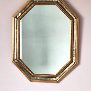 Op de afbeelding: Een achthoekige spiegel met een goudkleurige lijst. De spiegel heeft een decoratieve rand met een getextureerd, zilverkleurig accent. De spiegel hangt aan een lichtroze muur. De afmetingen van de spiegel zijn niet zichtbaar.