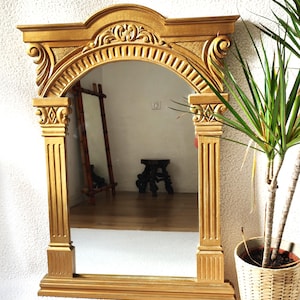 May include: A gold-colored, ornate mirror with an arched top and decorative columns. The mirror reflects a room with wooden floors and furniture. A potted plant sits to the right of the mirror.