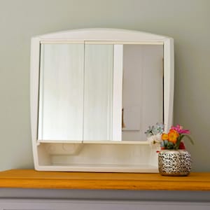 Peut inclure: Armoire à pharmacie blanche avec deux portes miroir et une étagère. L'armoire est fixée au mur au-dessus d'une commode avec un dessus en bois et un tiroir gris.