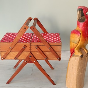 Può includere: Un portagioie in legno con un motivo a cuore rosso e bianco sul coperchio e una statua di pappagallo in legno intagliato con vernice rossa, gialla e arancione. Il portagioie ha un design pieghevole.
