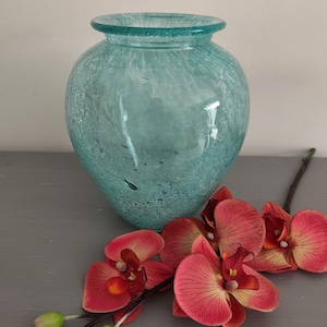May include: A bulbous, light turquoise glass vase with a textured, speckled finish and a wide, rounded rim. Beside the vase lies a stem of artificial orchid flowers featuring vibrant coral-pink petals with dark red centers and two small green buds.