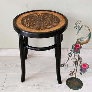 May include: A small, round wooden stool with a carved floral design on the seat. The seat is a light brown color, and the frame is black. A decorative metal peacock candle holder is next to the stool.