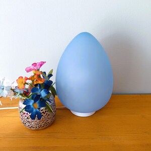 Peut inclure: Une lampe en forme d'œuf bleu clair avec une base blanche est posée sur une surface en bois à côté d'un vase argenté avec des fleurs artificielles colorées.
