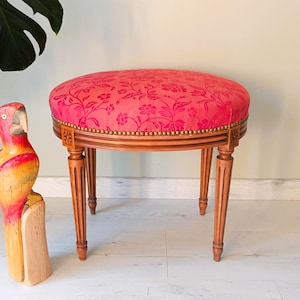 Puede incluir: Un pequeño taburete de estilo antiguo con un asiento redondo rojo con estampado floral. El marco de madera tiene cuatro patas delgadas y acanaladas y un ribete decorativo con pequeños tachuelas doradas. A la izquierda hay una estatua de loro tallada en madera.
