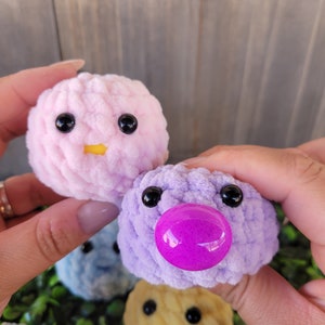 May include: Four crocheted plush toys in the shape of round balls with faces. The toys are pink, purple, blue, and yellow. The purple toy has a bright pink nose.