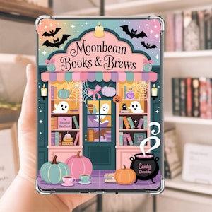 May include: A clear case with a colourful illustration of a bookstore named "Moonbeam Books & Brews." The design includes pumpkins, ghosts, bats, and a cauldron with the text "Candy Chronicles." The case is held in a person's hand.