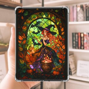 May include: A clear tablet case displaying a stained-glass style image of a witch in a black hat and purple dress. The witch is standing in front of a green window with bats, surrounded by orange and yellow autumn leaves. A cauldron with glowing orbs sits below.