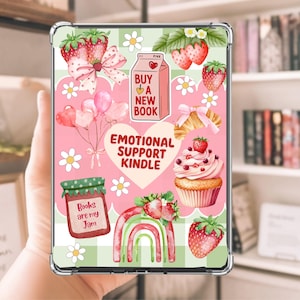 May include: A clear Kindle case with a pink and green design featuring strawberries, daisies, and the text "Buy a New Book" and "Emotional Support Kindle". Other graphics include a cupcake, a rainbow, and a jar of jam.