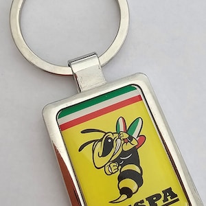 Könnte beinhalten: Ein silberner Schlüsselanhänger mit einem italienischen Flaggen-Design in Gelb, Grün und Rot. Der Schlüsselanhänger zeigt eine Cartoon-Biene mit dem Wort "VESPA" darunter.