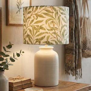 May include: A cream ceramic table lamp with a cylindrical base and a lampshade patterned with green leaves on a cream background. The lamp is on a wooden surface, with a small vase and framed botanical prints nearby.