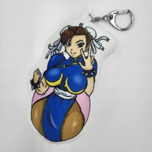 May include: A plush keychain featuring a cartoon character in a blue and yellow outfit with a white headband. The character has brown hair and is making a peace sign with her right hand.