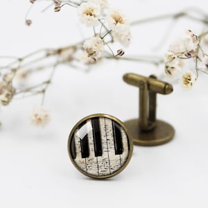 Può includere: Un paio di gemelli con un cabochon rotondo in vetro che presenta un design di spartito musicale vintage con tasti di pianoforte. I gemelli sono in ottone e hanno una finitura lucida.