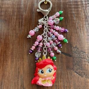 May include: A keychain featuring a cartoon figure of a princess with red hair and a gold crown. The keychain has pink, purple, and clear beads, and a silver clasp. The beads are in various sizes and shapes, with some having a sparkly appearance.
