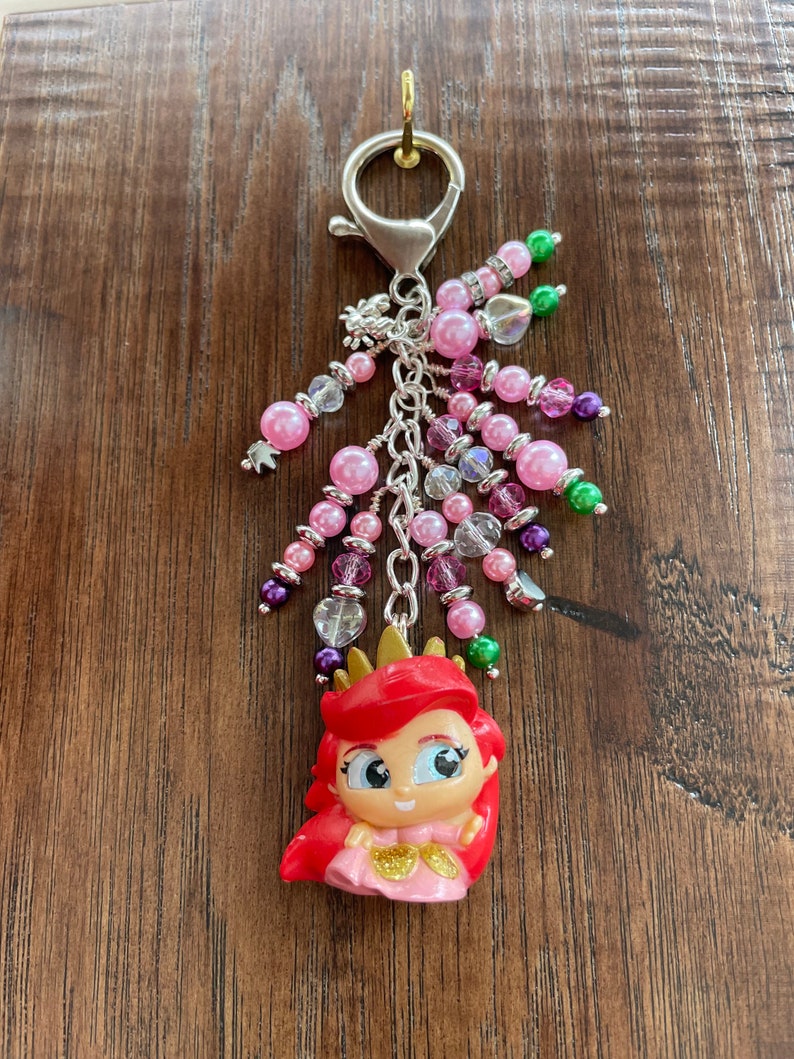May include: A keychain featuring a cartoon figure of a princess with red hair and a gold crown. The keychain has pink, purple, and clear beads, and a silver clasp. The beads are in various sizes and shapes, with some having a sparkly appearance.