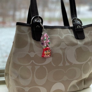 May include: A beige tote bag with a repeating logo pattern, black leather handles, and a decorative charm. The charm features a small figurine of a mermaid with pink hair, surrounded by pink and green beads.