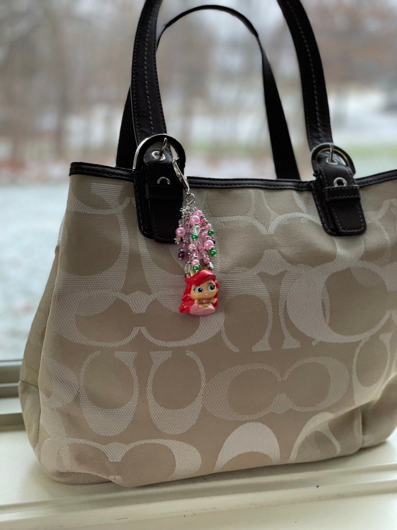 May include: A beige tote bag with a repeating logo pattern, black leather handles, and a decorative charm. The charm features a small figurine of a mermaid with pink hair, surrounded by pink and green beads.