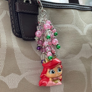 May include: Ariel-themed keychain charm with a cluster of pink and green beads. The charm features a small Ariel figurine with red hair, a pink dress, and a gold crown. The keychain is attached to a brown leather strap.