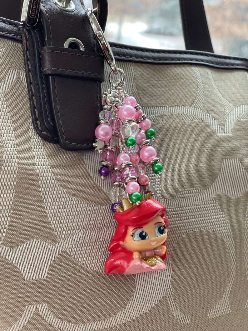May include: Ariel-themed keychain charm with a cluster of pink and green beads. The charm features a small Ariel figurine with red hair, a pink dress, and a gold crown. The keychain is attached to a brown leather strap.