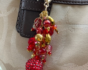 May include: A decorative bag charm with a gold-tone clasp. The charm features a cluster of red and gold beads, including a large, sparkling red sphere. The charm is attached to a brown leather strap on a beige handbag.