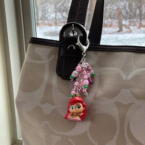 May include: Ariel-themed charm hanging from a purse. The charm features a small Ariel figurine with a red hair and pink dress, surrounded by pink, green, and purple beads. The purse is beige with a brown handle.