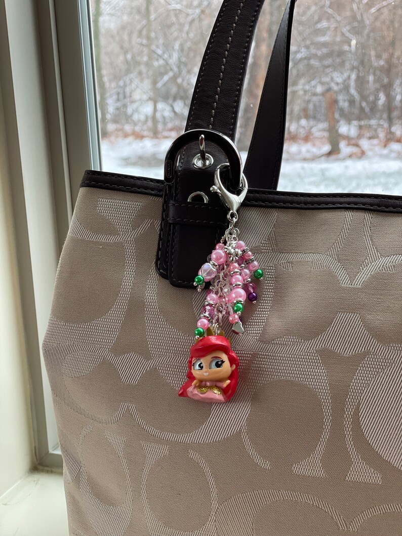 May include: Ariel-themed charm hanging from a purse. The charm features a small Ariel figurine with a red hair and pink dress, surrounded by pink, green, and purple beads. The purse is beige with a brown handle.
