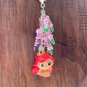 May include: A keychain charm featuring a cartoon character with red hair and a gold crown. The charm is adorned with pink, green, purple, and clear beads, and a silver-colored clasp. The background is a wooden surface.