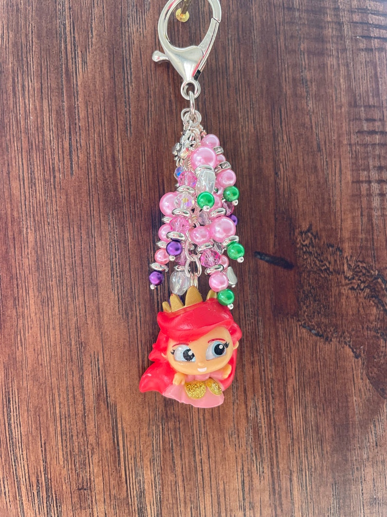 May include: A keychain charm featuring a cartoon character with red hair and a gold crown. The charm is adorned with pink, green, purple, and clear beads, and a silver-colored clasp. The background is a wooden surface.