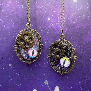 May include: Two oval pendants on bronze-toned chains, each featuring an ornate filigree frame, a metallic gear accent, and a central eye-shaped cabochon. One pendant displays a blue and white eye with purple glitter, while the other has a yellow and white eye.