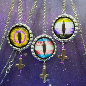 May include: Three silver-toned necklaces featuring bottle cap pendants with glass cabochons. Each pendant displays a vertical slit pupil eye design over different colored backgrounds, including yellow, pink, and flag patterns, with a small cross charm.