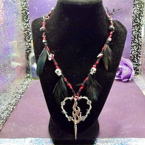 Pode incluir: Um colar de estilo gótico com uma corrente de contas pretas e vermelhas, adornado com penas pretas e pequenos pingentes de caveira. O pingente é um coração prateado com um design de chama e uma base pontiaguda.