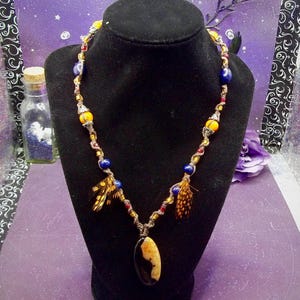 May include: A handcrafted necklace featuring a dark brown cord adorned with yellow, blue, and red beads. The necklace includes small feathers and a pendant with a black and tan oval stone. The necklace is displayed on a black mannequin.