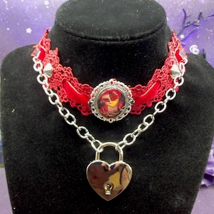 Könnte beinhalten: Ein rotes Spitzen-Choker-Halsband mit silberner Kette und herzförmigem Vorhängeschloss. Das Choker-Halsband hat ein zentrales Cameo und silberne Akzente. Die Halskette wird auf einer schwarzen Schaufensterpuppe präsentiert.