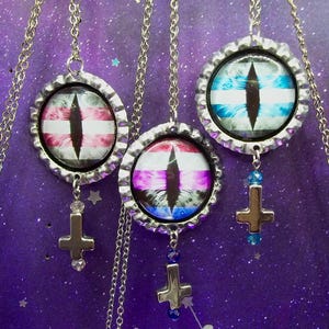May include: Three silver-toned metal bottle cap pendants hanging on thin chains. Each pendant features a circular graphic of a vertical slit pupil eye in pink, purple, or blue tones. A small silver cross charm hangs from the bottom of each pendant.