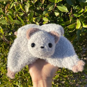 May include: A handmade, plush gray bat with brown ears and feet. The bat has large, black eyes and a small, stitched nose. The wings are spread, and the toy is held in a hand against a backdrop of green foliage.