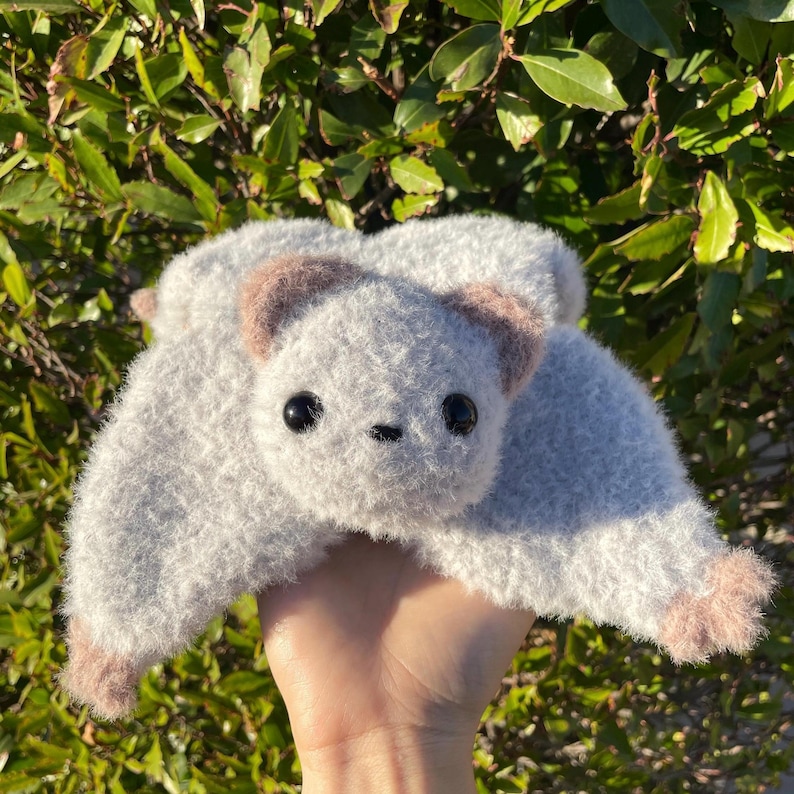 May include: A handmade, plush gray bat with brown ears and feet. The bat has large, black eyes and a small, stitched nose. The wings are spread, and the toy is held in a hand against a backdrop of green foliage.