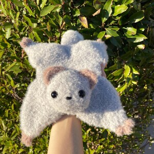 May include: A handmade, plush flying squirrel toy in soft gray with brown accents on the ears and feet. The toy has large, black eyes and a small, stitched nose, giving it a cute, friendly appearance. The plush is held against a backdrop of green foliage.