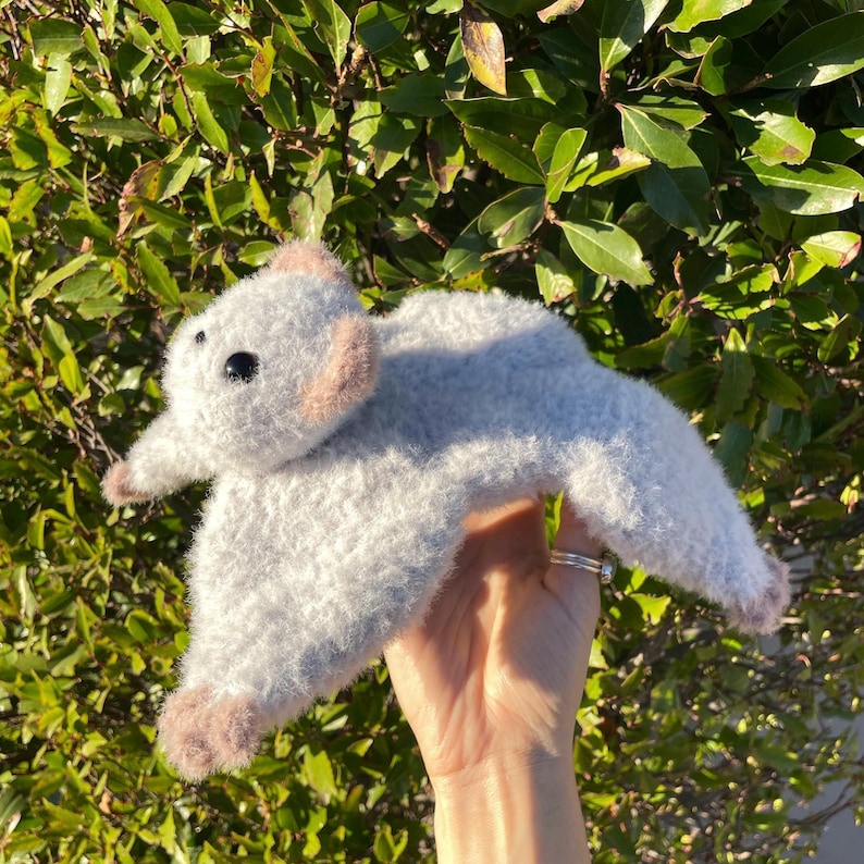 May include: A handmade, plush gray flying squirrel toy with light brown accents on the ears and feet. The toy has black eyes and a fluffy texture, perfect for a child's room or as a unique gift.