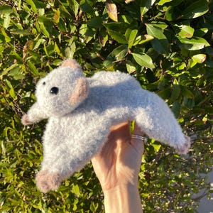 May include: A handmade, plush gray bat with black eyes and light brown accents on the ears and feet. The bat is held up against a backdrop of green foliage, showcasing its soft, textured appearance.