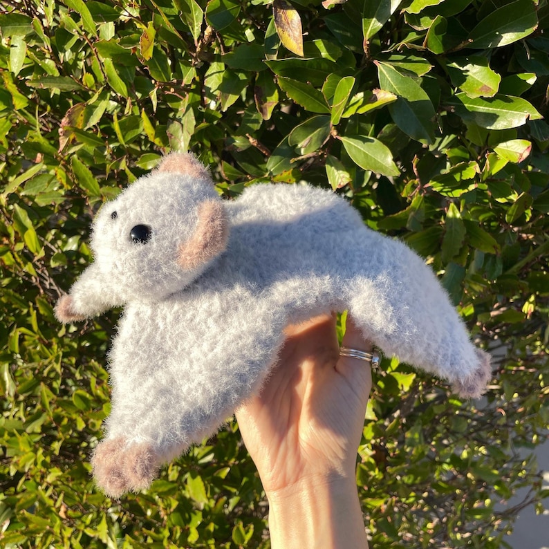 May include: A handmade, plush gray bat with black eyes and light brown accents on the ears and feet. The bat is held up against a backdrop of green foliage, showcasing its soft, textured appearance.
