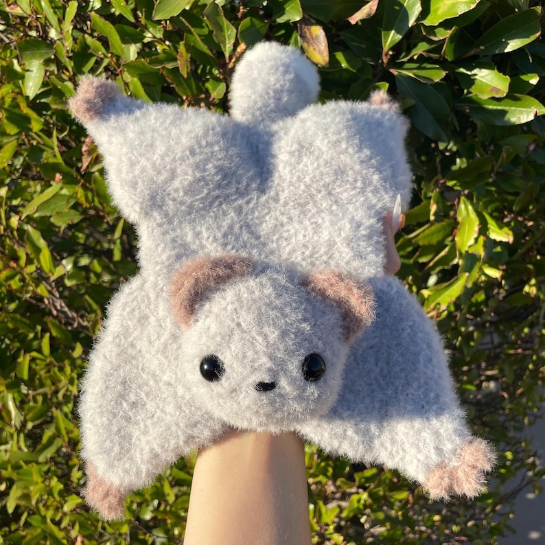 May include: A plush, gray flying squirrel hat with a soft, fuzzy texture. The hat features a detailed face with black eyes and brown accents on the ears and paws. The hat is designed to resemble a flying squirrel.