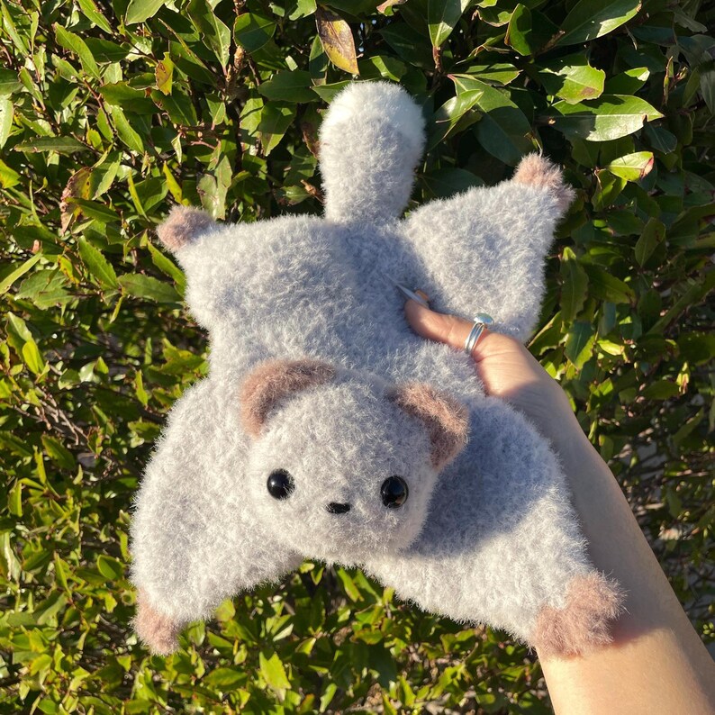 May include: A handmade gray plush toy in the shape of a flying squirrel. The toy has a fluffy texture, with brown accents on the ears and feet. It features black button eyes and a small, stitched nose. The toy is held against a backdrop of green foliage.