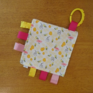 May include: A white baby sensory square with a bee and honeycomb pattern. It has four fabric ribbons in pink, yellow, and pink, and a yellow plastic teething ring attached to a pink ribbon.