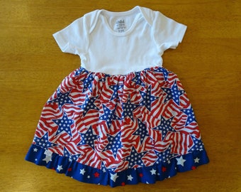 May include: A white onesie with short sleeves and a red, white, and blue American flag print skirt with stars. The onesie has the text "child of mine carter's 3-6M" printed on the front.